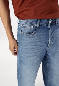 Personne portant un jean en denim bleu clair et une chemise couleur rouille rentrée dans le pantalon, avec la main posée près de la poche sur fond blanc.