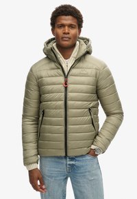 Uomo che indossa una giacca imbottita con cappuccio di un verde oliva chiaro sopra un maglione bianco sporco e jeans azzurri, in piedi con una mano in tasca.