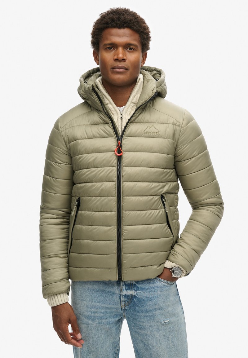 Mężczyzna w jasnooliwkowej pikowanej kurtce z kapturem na off-white swetrze i jasnoniebieskich dżinsach, stojący z jedną ręką w kieszeni.