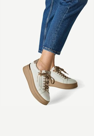 Person iført hvide og beige platformsneakers med brune snørebånd og blå jeans, der er cropsede over anklen.