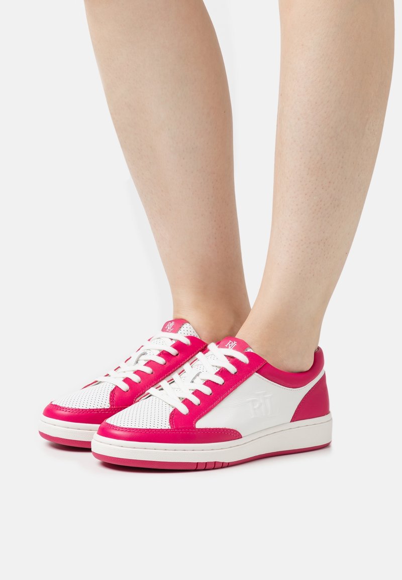 Personne portant des baskets blanches avec des accents rose vif, des lacets blancs et un bout perforé, debout sur un fond blanc uni.