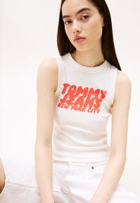 Valge ilma varrukateta top, millel on ribakujuline tekstuur ja ees julge punane "TOMMY JEANS NEW YORK CITY" graafika, paari heledate pükstega.