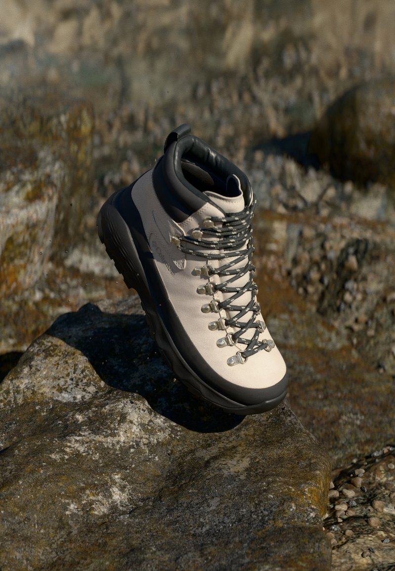 Bota de caminhada de um bege claro com sola de borracha preta, parte superior flexível, ilhós de metal e atacadores cinzentos, posicionada sobre uma superfície rochosa.