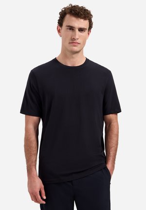 Jonge man met krullend haar, gekleed in een effen zwart T-shirt met korte mouwen en een donkere broek, staand tegen een witte achtergrond.