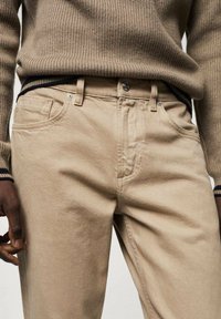 Beige Baumwollhosen mit klassischer Passform, ausgestattet mit einem Reißverschluss, Fronttaschen und einer subtilen Textur. Abgerundet durch einen blau gestreiften Bund.