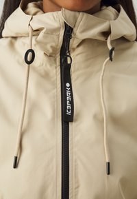 Béžová bunda s kapucňou s čiernym zipsom a výťahovým jazdcom, akcentmi s šnúrkou a hladkou, matnou textúrou. Logo značky zobrazené na zips.