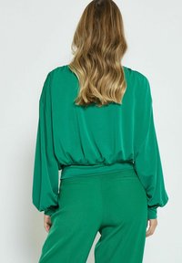 Top verde a maniche lunghe con orlo arricciato, realizzato in tessuto liscio. Presenta una vestibilità ampia e maniche lievemente a sbuffo, abbinato a pantaloni coordinati.