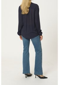 Femme aux longs cheveux blonds portant un chemisier bleu marine, un jean bootcut bleu et des chaussures slingback à talons noirs à bout pointu, debout devant un fond uni.