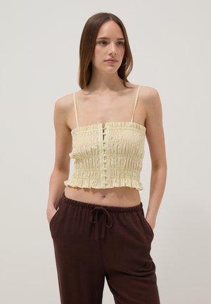 Jeune femme aux cheveux bruns raides porte un top court smocké jaune pâle et un pantalon marron foncé avec cordon, debout les mains dans les poches.