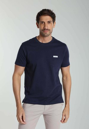 Homme aux cheveux courts et foncés portant un t-shirt bleu marine à manches courtes et un pantalon beige clair sur un fond clair uni.
