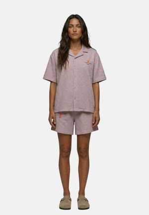 Femme aux longs cheveux foncés portant une chemise texturée violet clair à manches courtes avec boutons et un short assorti, debout les bras le long du corps sur fond blanc.