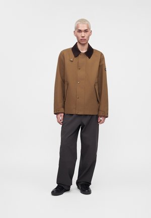 Un mannequin se tient devant un fond blanc uni, vêtu d’un manteau marron à col foncé, d’un pantalon noir ample et de chaussures noires.