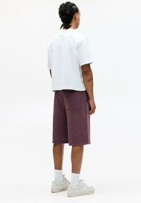 T-shirt en coton blanc avec un design court, associé à un short violet avec une poche arrière et un ourlet côtelé, porté avec des baskets blanches.