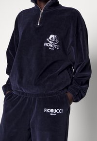Personne portant un survêtement en velours Fiorucci Milan bleu marine foncé avec broderie de logo blanc sur la poitrine et la cuisse, les mains dans les poches.