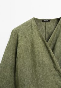 Grünes Leinen-Wrap-Top mit V-Ausschnitt, ausgestattet mit einer strukturierten Oberfläche und verstärkten Nähten, entworfen für einen lässigen Sitz. Markentag sichtbar.