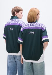 Πράσινα και μοβ σακάκια με κοντά μανίκια και γεωμετρικό μοτίβο στο πάνω μέρος, με λευκή λεπτομέρεια στο τελείωμα και κεντημένο λογότυπο.