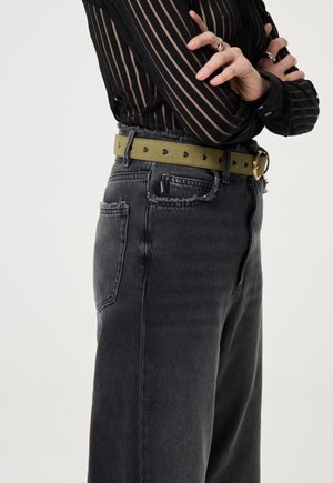 Hoge taille zwarte denim jeans met rafelige randen, gecombineerd met een groene riem met zwarte hartvormige uitsparingen. Doorzichtige top met verticale strepen.