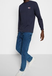 Marinblå långärmad T-shirt med "TOMMY JEANS"-logotyp, i kombination med blå denimjeans och vita sneakers. Avslappnad outfit med enkla linjer och passform.