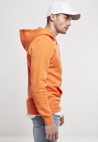 Sudadera naranja con capucha ajustable, puños y dobladillo de canalé, tejido suave, combinada con jeans azul claro y una gorra blanca.
