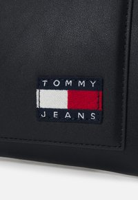 Zwarte leren tas met een rechthoekige patch met de tekst "TOMMY JEANS" en een logo ontwerp in rood, wit en blauw. Glad oppervlak.