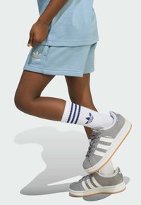 Grå mocka sneakers med tre vita ränder, i kombination med vita strumpor med blå ränder och en blå logotyp, samt ljusblå shorts.