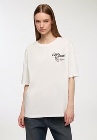 Biała, oversize'owa koszulka z czarnym nadrukiem tekstu "Ciao Amore!" oraz wzorem serca na lewej stronie klatki piersiowej. Materiał bawełniany, krótki rękaw.