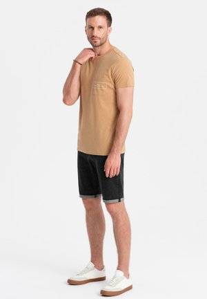 Beige Baumwoll-T-Shirt mit Brusttasche, kombiniert mit dunkelblauen Jeansshorts und weißen Sneakers mit braunen Sohlen.