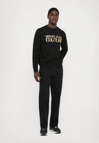 Črna sweats jednodavna s zlatim logotipom "VERSACE JEANS COUTURE", v kombinaciji z črnimi hlačami z belimi šivi, obuti v črne superge.