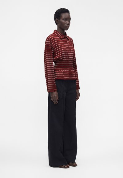 Femme debout, portant un haut rayé rouge et blanc à manches longues avec taille froncée et un pantalon noir à jambes larges sur un fond blanc uni.