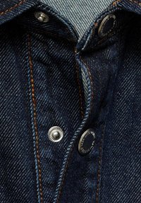 Tela de mezclilla oscura con costuras naranjas contrastantes; cuenta con botones de presión de metal y un diseño de cuello clásico. La vista en primer plano muestra la textura.