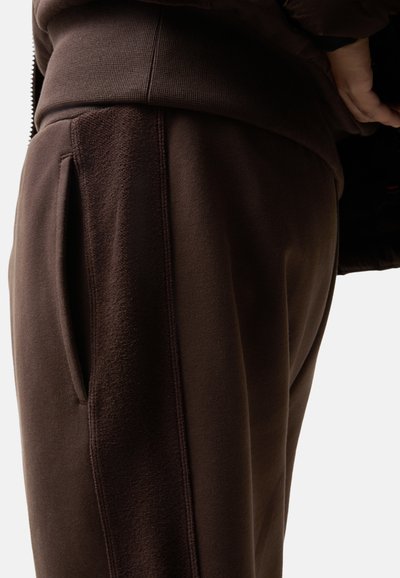 Brune joggers lavet af blødt, struktureret stof med en sidelomme og synlige syninger. Tæt fit, der viser en afdampet form.