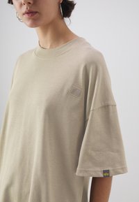 Beige bavlněné tričko s krátkými rukávy, kulatým výstřihem a malou látkovou nášivkou na hrudi a na lemu rukávu. Hladká textura.