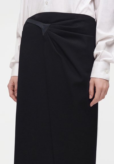 Femme portant une jupe noire taille haute avec un détail drapé et une blouse blanche boutonnée à manches longues.