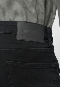 Jeans in denim nero con una vita di tessuto grigio scuro testurizzato. Un'etichetta rettangolare in pelle sul retro mostra il nome del marchio "ONLY & SONS".