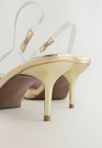 Zlaté sandále na vysokom opätku s priehľadnými pásikmi. Elegantný dizajn s špicatými prstami, tenkými členkovými pásikmi a stiletto opätkami s lesklým povrchom.