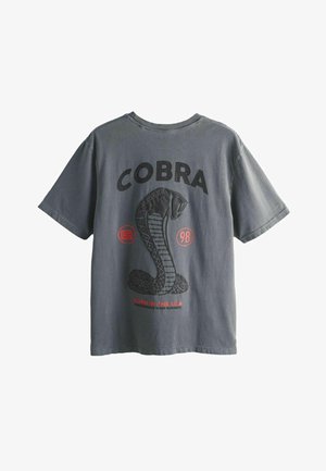 Tumehall shirt musta groter musta kobragraafika ja tekstiga "COBRA" ülalt, punaste aktsentidega, sealhulgas tekstiga "Born in the USA" all.