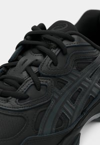 Gros plan d'une chaussure de sport noire avec un tissu texturé, des surcouches synthétiques et des lacets noirs noués en nœud sur fond blanc.