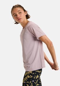 Young woman stretching arms behind back, wearing a light purple short-sleeve shirt and black leggings with yellow floral patterns.