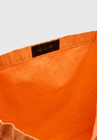 Orange tygväska inuti med mjuk textur, med en svart etikett högst upp som lyder "ALLSAINTS." Ingen synlig hårdvara eller mönster.