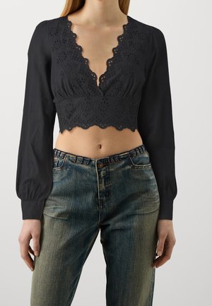 Femme portant un chemisier court noir à manches longues avec une dentelle à œillets festonnée et un jean bleu taille haute avec des boutons et des détails à œillets.