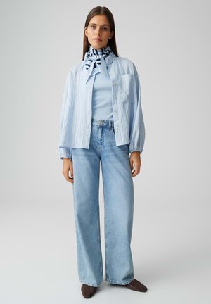 Giovane donna che indossa una camicetta azzurro chiaro, sciarpa a righe al collo, jeans a gamba larga e scarpe scure con punta, in piedi su uno sfondo uniforme.