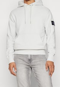 Vit hoodie gjord av mjukt material med en framficka, snoddar och en logopatch på vänster ärm. Har ribbade muddar och fåll.