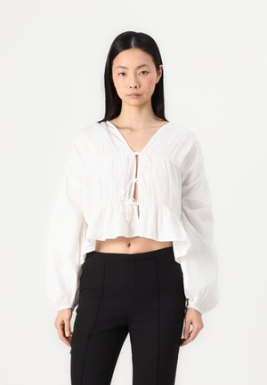 White cropped blouse with a V-neckline and drawstring front; features long puff sleeves and a ruffled hem. Paired with black tailored pants.