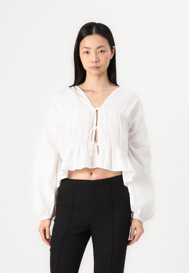 Blusa bianca corta con scollatura a V e davanti con cordoncino; presenta maniche lunghe a palloncino e orlo arricciato. Abbinata a pantaloni neri su misura.