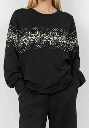 Mujer con un suéter negro de manga larga con un patrón de copos de nieve tejidos en color crema en el pecho y las mangas, con las manos en los bolsillos.