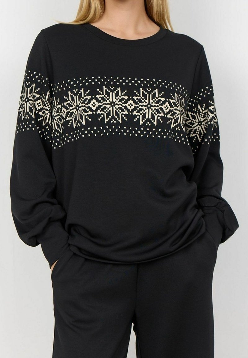 Femme portant un haut noir à manches longues avec un motif flocon de neige en tricot couleur crème sur la poitrine et les manches, les mains dans les poches.