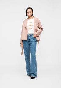 Γυναίκα που στέκεται φορώντας ένα μπεζ ανοιχτό σακάκι, λευκό μπλουζάκι με σχέδιο φτερών, μπλε τζιν με φαρδιά μπατζάκια και μαύρα παπούτσια με μυτερή μύτη.