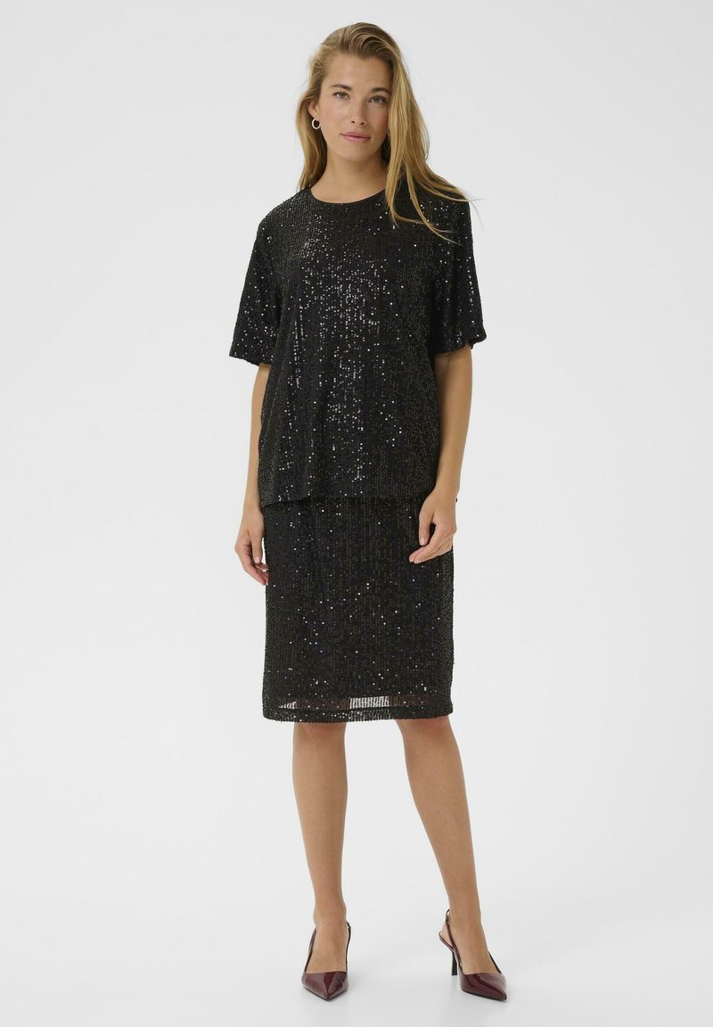 Vrouw in een kort zwart glitst shirt met korte mouwen en een bijpassende rok tot de knie, staande met maroon hakken tegen een effen witte achtergrond.