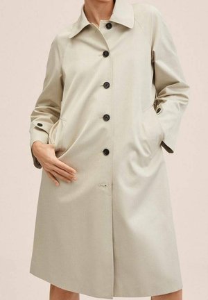 Beige knie-lange jas met zwarte knopen, lange mouwen, kraag en twee zijzakken, gedragen met één hand in de zak en één hand rustend op de jas.