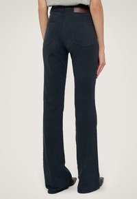 Pantalones oscuros de corte acampanado con una textura suave, bolsillos traseros y un parche de cuero en la cintura. Terminado con detalles de costura sutiles.